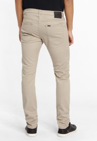 Man som bär beige slim-fit jeans och svarta sneakers, står vänd bort med händerna avslappnat vid sidorna på vit bakgrund.