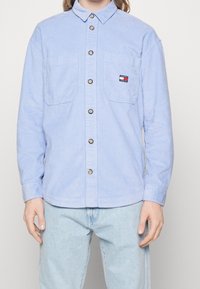 Jasnoniebieska koszula ze sztruksu w paski, z kołnierzykiem zapinanym na guziki, przednimi kieszeniami i naszywką z logo przy lewej kieszeni. Guziki z przodu.