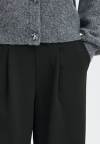 Cardigan en tricot gris avec des boutons décoratifs bleus à motifs floraux associé à un pantalon noir ample.