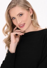 Czarny sweter o teksturowanej fakturze z szerokim dekoltem. Modelka nosi okrągłe złote kolczyki z białymi perłami, ma miękkie, falujące blond włosy.