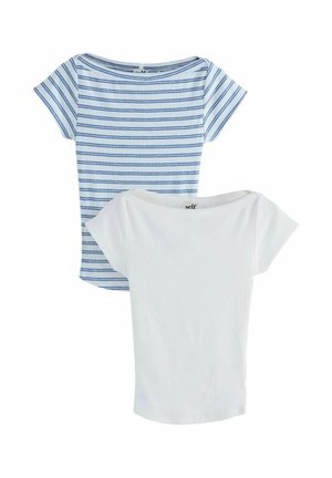 Junge Frau mit lockigem Haar, die ein weiß-blau gestreiftes Crop-Top und weiße Hosen trägt, steht mit einer Hand in der Tasche.