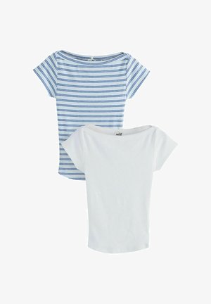 Junge Frau mit lockigem Haar, die ein weiß-blau gestreiftes Crop-Top und weiße Hosen trägt, steht mit einer Hand in der Tasche.