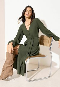 Olijfgroene, geribbelde jurk met lange mouwen, een V-hals, knoopdetails en een ceintuur in de taille. Model draagt beige suède kniehoge laarzen en zit op een stoel.