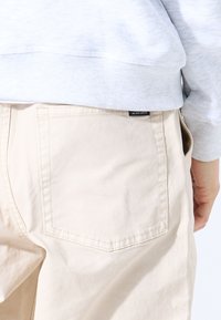 Pantalones de algodón beige con un bolsillo trasero que lleva una etiqueta visible con la inscripción "WE ARE GAEA". La tela parece suave con mínima textura.