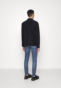 Camisa de ante negro con una yugo trasero y mangas largas, combinada con jeans de mezclilla azul y zapatos negros con cordones. Diseño simple, sin patrones visibles.