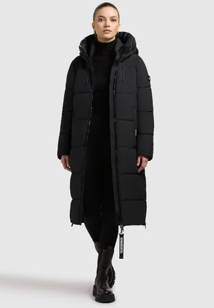 Sort pufferjakke med langt design, quiltet tekstur og hætte. Har en fuld lynlås og logo-tag ved kanten. Bæres over en sort turtleneck.