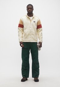 Άνδρας που φοράει μπεζ φλις μπουφάν με κόκκινες και καφέ ρίγες, σκούρο πράσινο παντελόνι, παπούτσια πεζοπορίας, κρατώντας ένα λευκό μπουκάλι νερού, στέκεται μπροστά σε λευκό φόντο.