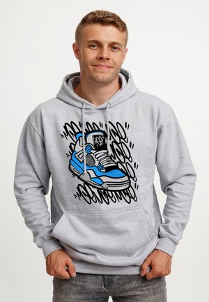Młody mężczyzna w jasnoszarej bluzie z kapturem z dużą niebiesko-czarną grafiką buta sportowego i abstrakcyjnymi czarnymi zygzakami na przodzie.