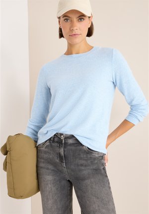 Lichtblauwe trui van zacht materiaal, ronde halslijn, opgerolde mouwen. Gecombineerd met donkergrijze jeans en een olijfgroene pufferjack vastgehouden.