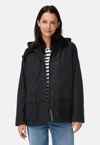 Vrouw met donker haar die een zwarte hoodie-jack over een gestreept shirt en blauwe jeans draagt, staande tegen een effen lichte achtergrond.