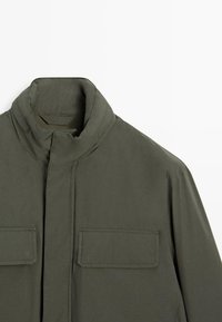 Giacca verde oliva con collo alto, due tasche anteriori e una texture liscia e opaca. Presenta un design minimalista e linee pulite.