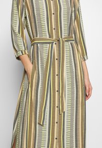 Femme portant une robe boutonnée ceinturée avec un motif à rayures verticales dans des tons de jaune, noir et beige, la main dans la poche.