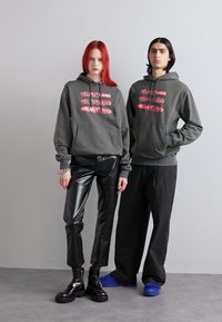 Grå hettegensere med "HELMUT LANG" trykt i rosa, parret med svarte lærbukser og avslappede mørke bukser. Svarte plattformstøvler og blå sko.