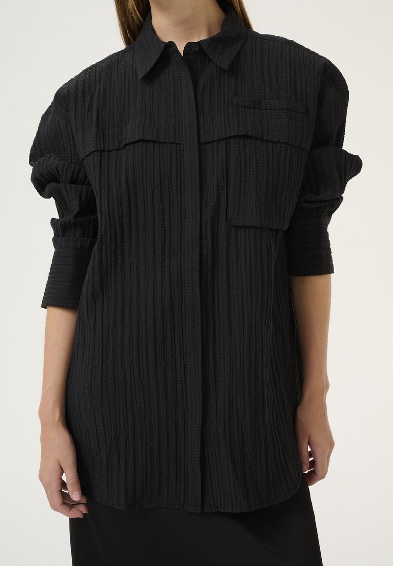 Camicia nera a maniche lunghe con un tessuto testurizzato a coste, dotata di due tasche sul petto e dettagli arricciati sulle maniche.
