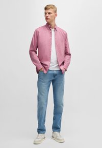 Hele roosa nööbitav särk valge t-särgi peal, kombineeritud helesiniste teksade ja beežide tume triibuliste tennistega, seistes neutraalse tausta ees.