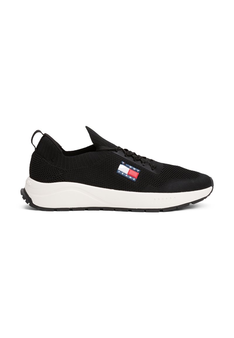 Baskets de sport noires avec un dessus en mesh respirant, semelle intermédiaire blanche, semelle extérieure en caoutchouc texturé, et un petit logo rouge, blanc et bleu sur le côté.