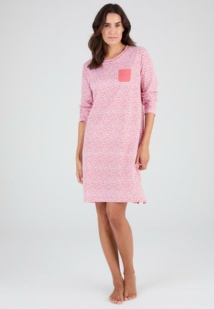 Donna in piedi a piedi nudi che indossa una camicia da notte rosa e bianca con stampa leopardata, maniche lunghe e una tasca sul petto di colore rosa solido.