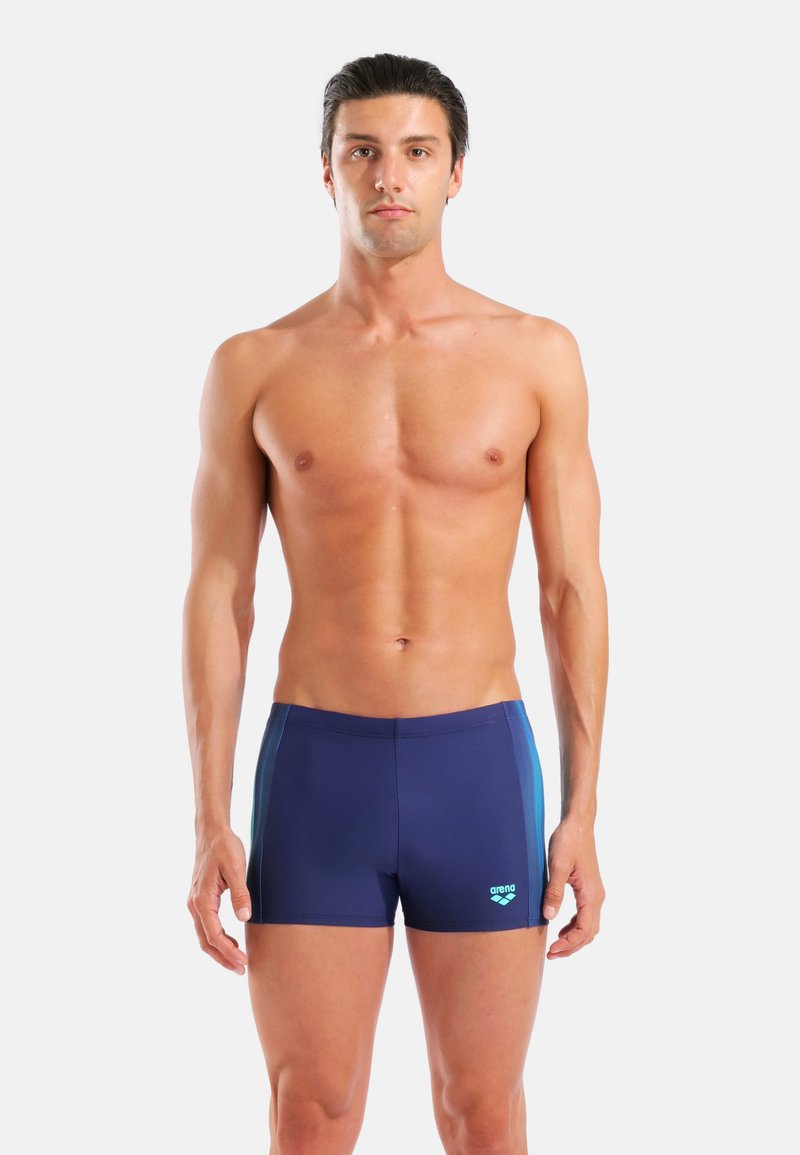 Bañador de hombre en azul marino con paneles laterales en verde azulado. Hecho de un material elástico y de secado rápido, con un logo de arena en la parte delantera.