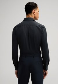 Camicia nera a maniche lunghe con una trama liscia e colletto classico, caratterizzata da polsini con bottoni e una vestibilità su misura. Indossata con pantaloni scuri.