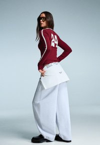 Bordeauxrotes Langarmshirt mit weißen Streifen und der Nummer 24. lockere graue Sweatpants, schwarze Schuhe und ein weißes Beuteltaschen-Accessoire um die Taille.