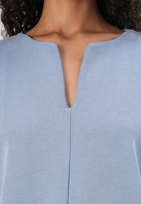 Top azul claro com decote em V, apresentando uma textura suave e uma costura central que desce verticalmente pela frente.