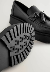 Mocasines de cuero negro con una parte superior texturizada, detalle de borlas y suela de goma gruesa que presenta un diseño de banda de rodadura para mayor agarre.