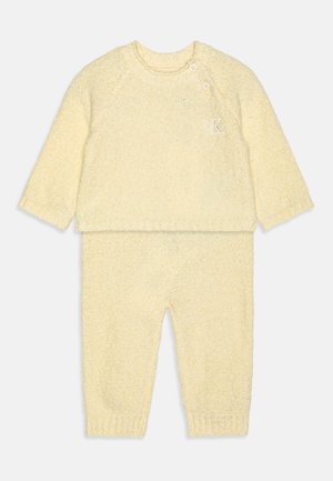 Rumena pletena dvodelna obleka, ki vključuje dolg rokav s krožnim izrezom in dvema gumboma ter usklajene hlače z elastičnimi manšetami.