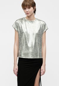 Νεαρό άτομο με κοντό μαλλί που φοράει ένα λαμπερό ασημένιο μπλουζάκι κοντομάνικο και μια μαύρη φούστα με ψηλή σχισμή, στέκεται μπροστά σε απλό φόντο.