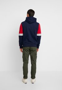 Felpa con cappuccio blu marina, maniche a strisce rosse e grigie, abbinata a pantaloni cargo oliva. Il tessuto appare morbido e informale.