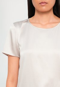 Giovane che indossa una blusa di raso beige chiaro a maniche corte, ripresa dal collo fino alla parte superiore del braccio su uno sfondo chiaro e semplice.