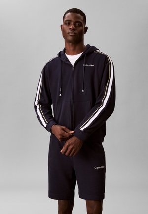 Άνδρας που φοράει μαύρο φούτερ με φερμουάρ Calvin Klein με λευκές και κίτρινες ρίγες στα μανίκια και ταιριαστά μαύρα σορτς, σε γκρι φόντο.