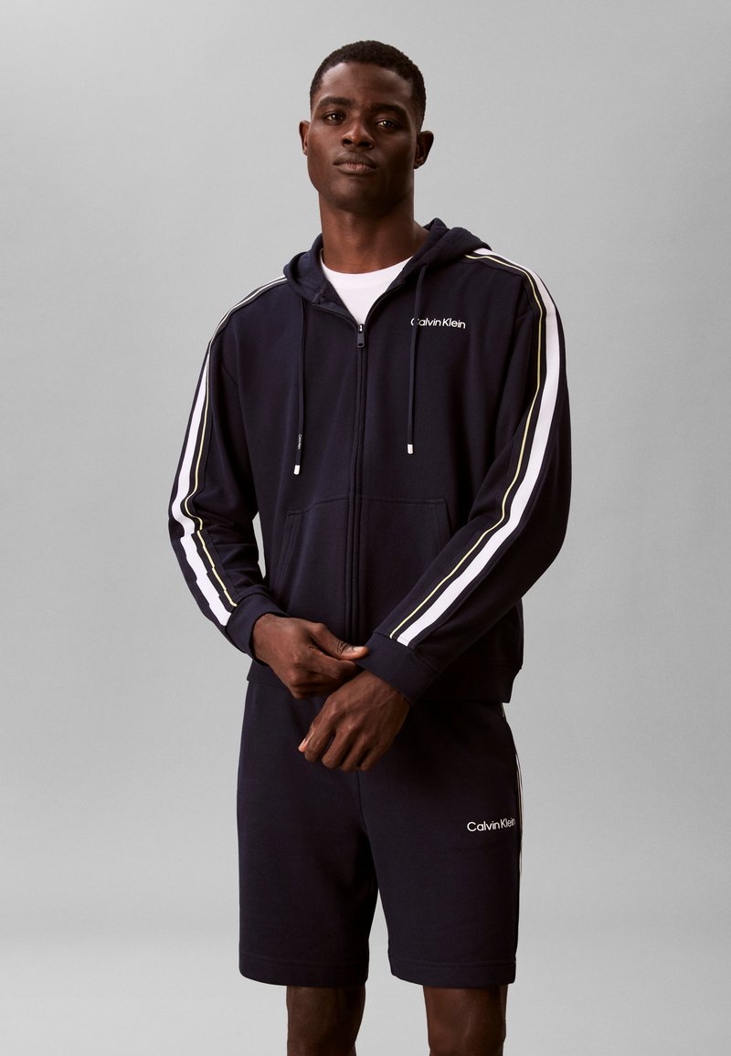 Άνδρας που φοράει μαύρο φούτερ με φερμουάρ Calvin Klein με λευκές και κίτρινες ρίγες στα μανίκια και ταιριαστά μαύρα σορτς, σε γκρι φόντο.