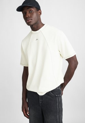 Cream-colored short-sleeve shirt with a ribbed crew neck, featuring raglan sleeves and a small logo at the chest. Dark jeans complement the outfit.