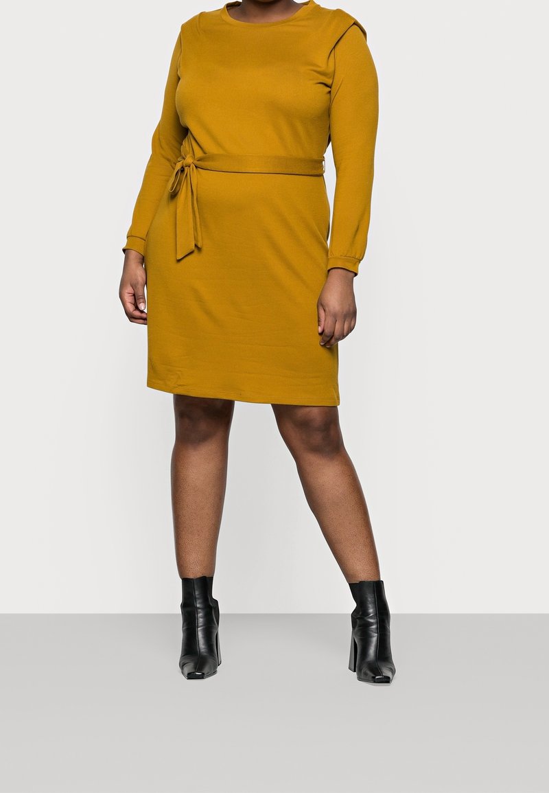 Robe jaune moutarde à manches longues avec un col rond, taille ceinturée et fentes latérales, associée à des bottines noires à la cheville. Texture lisse.