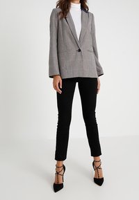 Šedý a bílý kostičkovaný blazer s klopami, dvěma předními kapsami, nošený přes bílý rolák, doplněný černými kalhotami a sandály na podpatku.