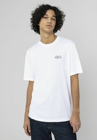 T-shirt en coton blanc avec manches courtes et col rond. Logo texte "Meet me Playground" en noir sur la partie gauche de la poitrine.