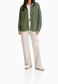 Grön bomullskavaj med två framfickor, tillsammans med ljusbeige cargo-byxor med sidofickor, och vita sneakers med bruna sulor.