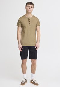 Maglietta henley a maniche corte verde oliva con quattro bottoni marroni. Abbinata a pantaloni corti neri e sneakers marroni con lacci bianchi e calze.