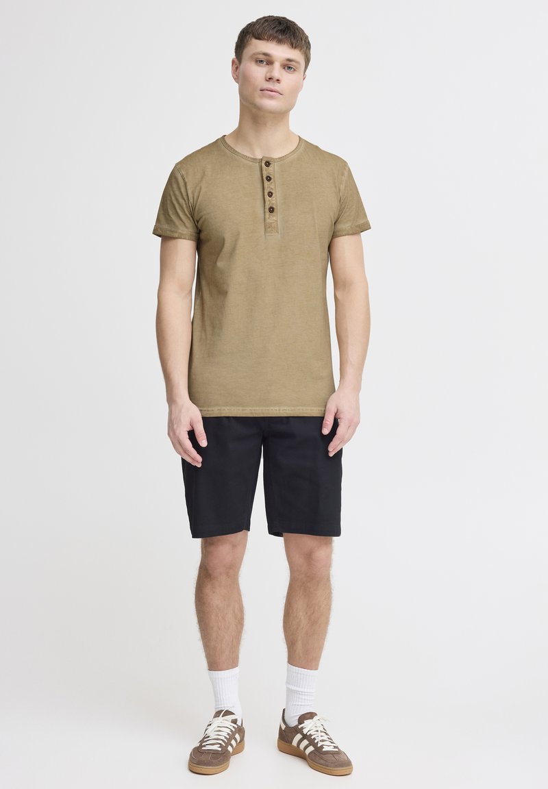 Maglietta henley a maniche corte verde oliva con quattro bottoni marroni. Abbinata a pantaloni corti neri e sneakers marroni con lacci bianchi e calze.