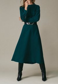 Donna che indossa un abito midi a maniche lunghe di un blu verde scuro con una cintura nera e stivali neri fino al ginocchio, in piedi davanti a uno sfondo semplice.
