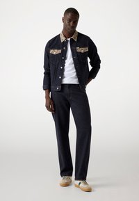 Dark denim jacket with patterned collar and pockets; paired with matching dark denim pants and beige sneakers featuring green accents.
