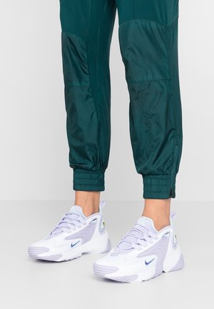 Hvide og lavendel Nike sneakers brugt med mørkegrønne bukser med elastiske manchetter, vist fra anklerne og nedad på en lysegrå gulv.