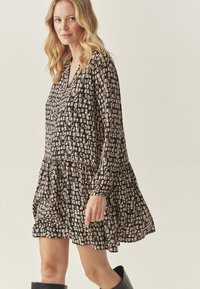 Juoda suknelė su geometriniu béžiniu raštu, turinti V formos iškirptę, ilgomis rankovėmis ir tiered sijonu. Pagaminta iš lengvos medžiagos.