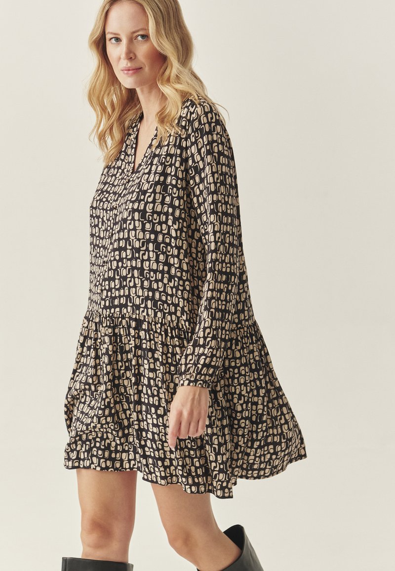 Juoda suknelė su geometriniu béžiniu raštu, turinti V formos iškirptę, ilgomis rankovėmis ir tiered sijonu. Pagaminta iš lengvos medžiagos.