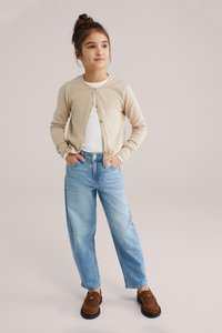 Noor tüdruk seisab käed taskutes, tal on seljas beež kardigan, valge särk, helesinised teksad värviliste plekidega ja pruunid loafersid.