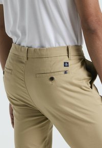 Pantalons kaki en tissu léger, dotés de deux poches arrière, d'une fermeture à bouton et d'un patch logo sur la ceinture.