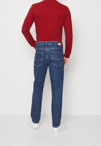 Mörkblå denimjeans med rak skärning, med bakfickor och en märkeslapp vid midjan, parat med en röd långärmad skjorta.