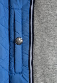 Primo piano di una tasca di una giacca trapuntata blu con un bottone d'argento incisa "MGD" accanto a un tessuto grigio bordato da una rifinitura a righe nere e bianche.