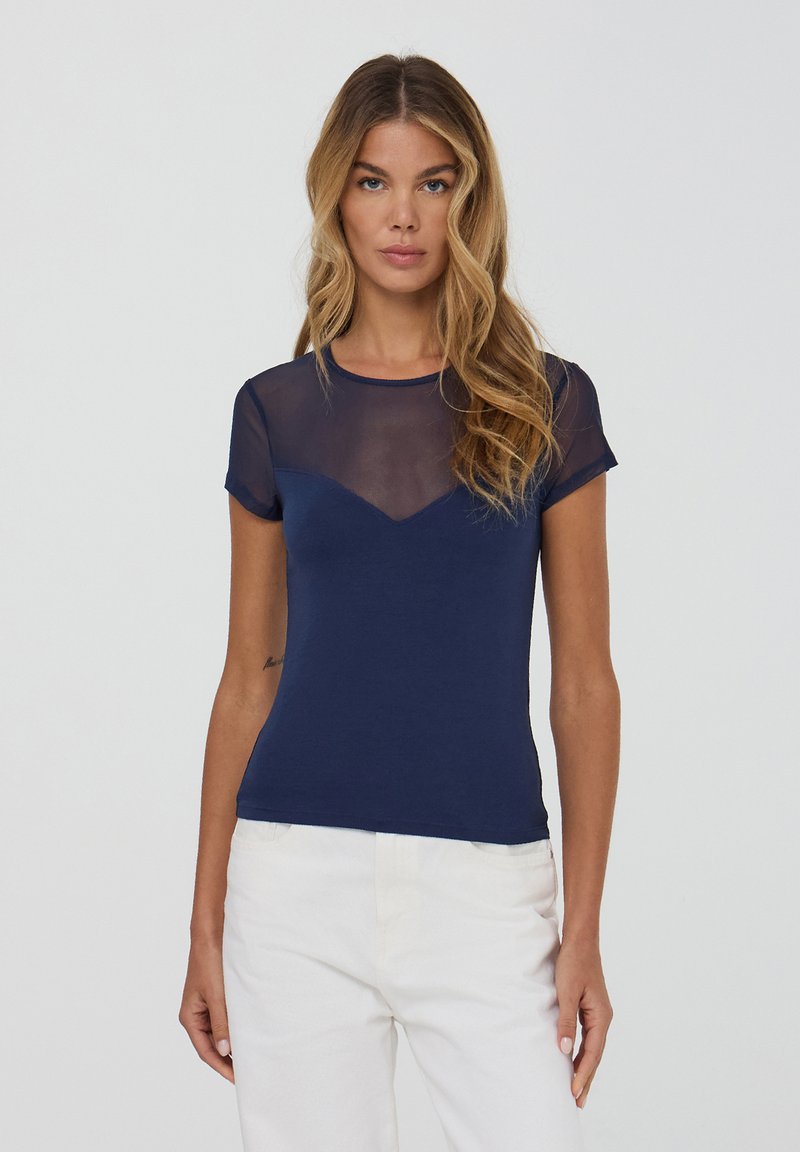 Donna con lunghi capelli mossi biondi indossa una maglietta blu navy a maniche corte con pannello trasparente superiore e pantaloni bianchi, in piedi davanti a uno sfondo semplice.