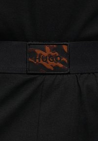 Tissu noir avec une étiquette camouflage noire et marron cousue au centre indiquant "HUGO" en lettres en relief.